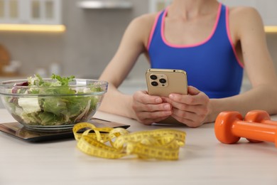 Photo of Woman using smartphone at white table with kitchen scale, salad, measuring tape and dumbbells indoors, closeup