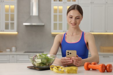 Photo of Woman using smartphone at white table with kitchen scale, salad, measuring tape and dumbbells indoors, space for text