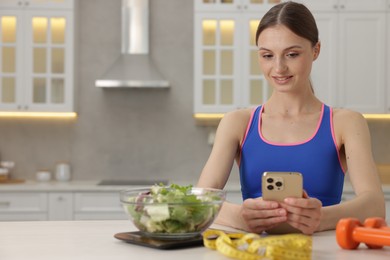 Photo of Woman using smartphone at white table with kitchen scale, salad, measuring tape and dumbbells indoors, space for text