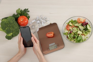 Photo of Woman using smartphone at white wooden table with kitchen scale, healthy food and measuring tape, top view
