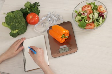 Photo of Woman developing diet plan at white wooden table with kitchen scale, healthy food and measuring tape, top view