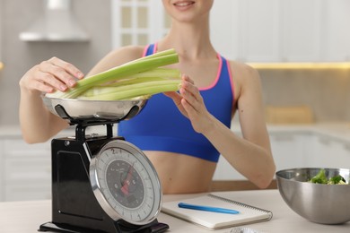 Photo of Woman weighting celery on kitchen scale at white table indoors, closeup