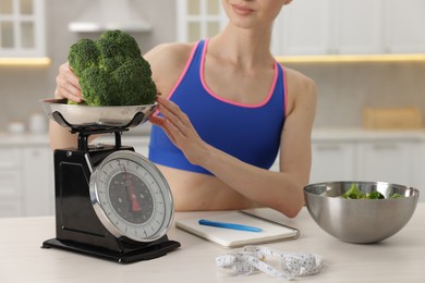 Photo of Woman weighting broccoli on kitchen scale at white table indoors, closeup
