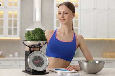 Photo of Woman weighting broccoli on kitchen scale at white table indoors