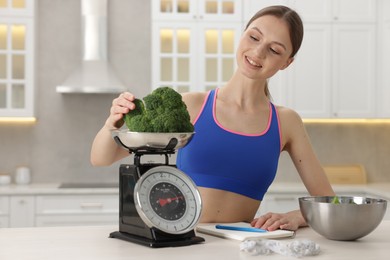Photo of Woman weighting broccoli on kitchen scale at white table indoors