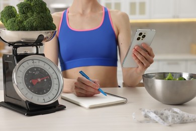 Photo of Woman with smartphone developing diet plan at white wooden table indoors, closeup