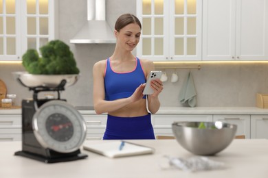 Photo of Woman using smartphone near table with kitchen scale, broccoli, notebook, measuring tape and salad indoors, selective focus