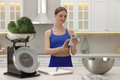 Photo of Woman using smartphone near table with kitchen scale, broccoli, notebook, measuring tape and salad indoors, selective focus