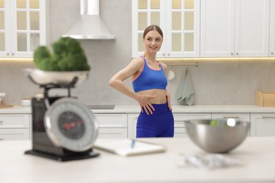 Photo of Woman posing near table with kitchen scale, broccoli, notebook, measuring tape and salad indoors, selective focus