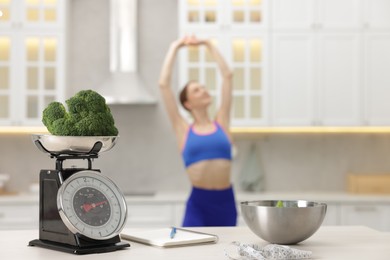 Photo of Slim woman posing indoors, focus on kitchen scale, broccoli, notebook, measuring tape and salad