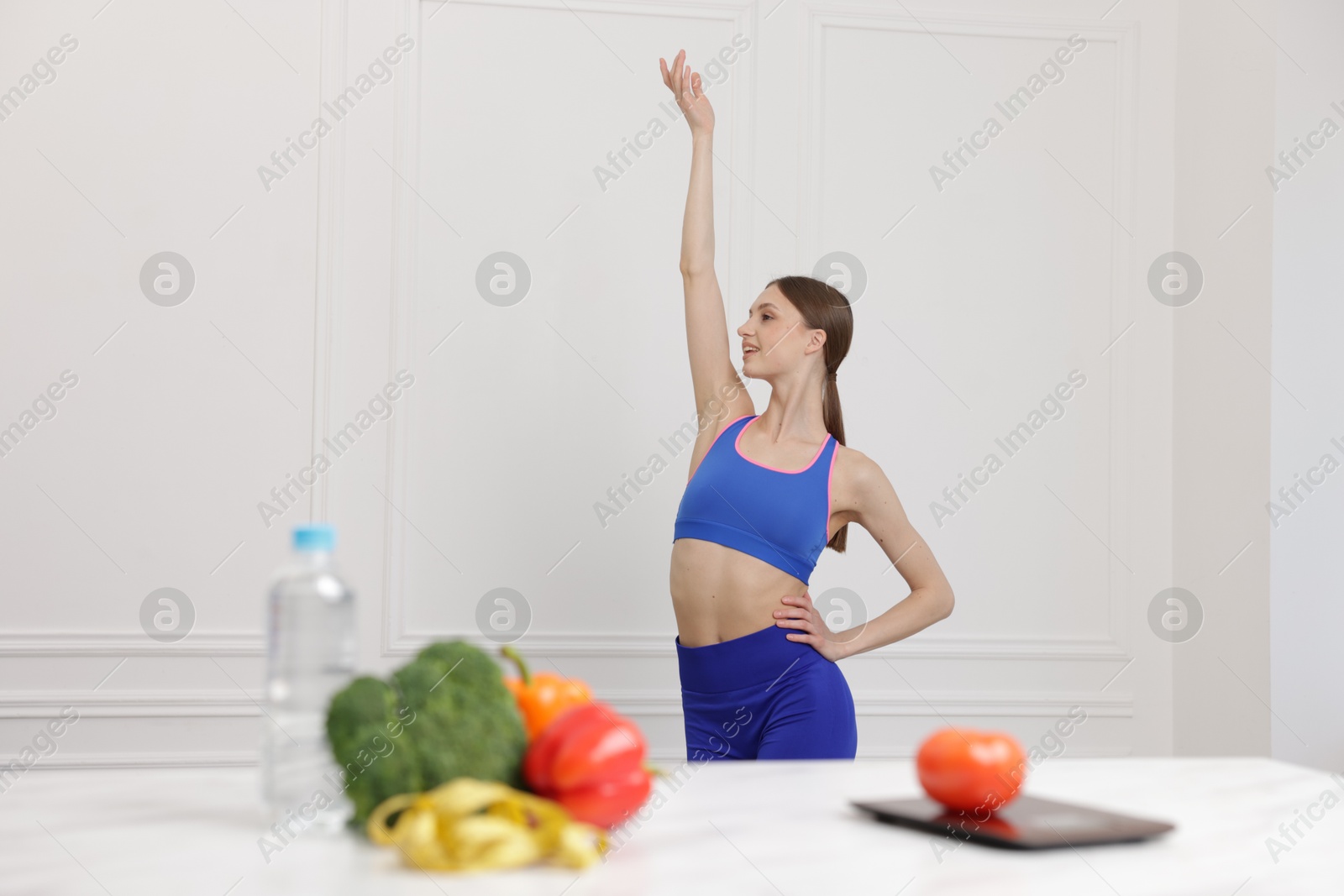 Slim woman posing near table with vegetables, water, measuring tape and kitchen scale indoors, selective focus Photo of Slim woman posing near table with vegetables, water, measuring tape and kitchen scale indoors, selective focus