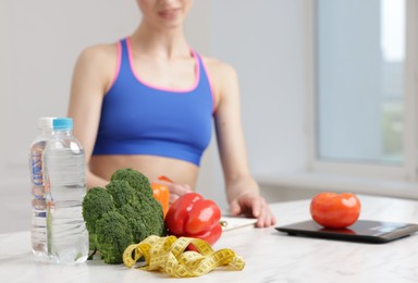 Photo of Woman developing diet plan at white marble table indoors, focus on vegetables, water and measuring tape, closeup