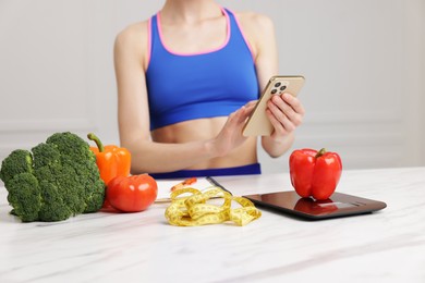 Photo of Woman using smartphone at white marble table with kitchen scale, vegetables and measuring tape indoors, closeup