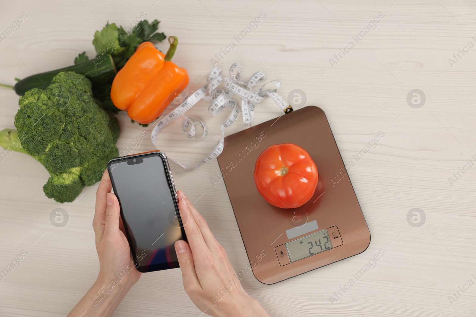 Photo of Woman using smartphone at white wooden table with kitchen scale, vegetables and measuring tape, top view