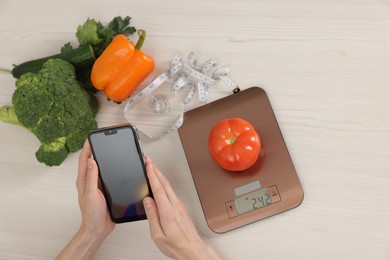 Photo of Woman using smartphone at white wooden table with kitchen scale, vegetables and measuring tape, top view