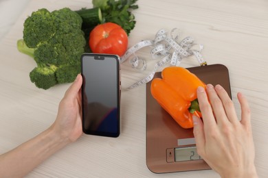 Photo of Woman with smartphone weighting bell pepper on kitchen scale at white wooden table, above view