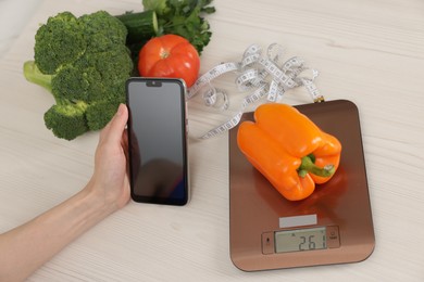 Photo of Woman using smartphone at white wooden table with kitchen scale, vegetables and measuring tape, above view