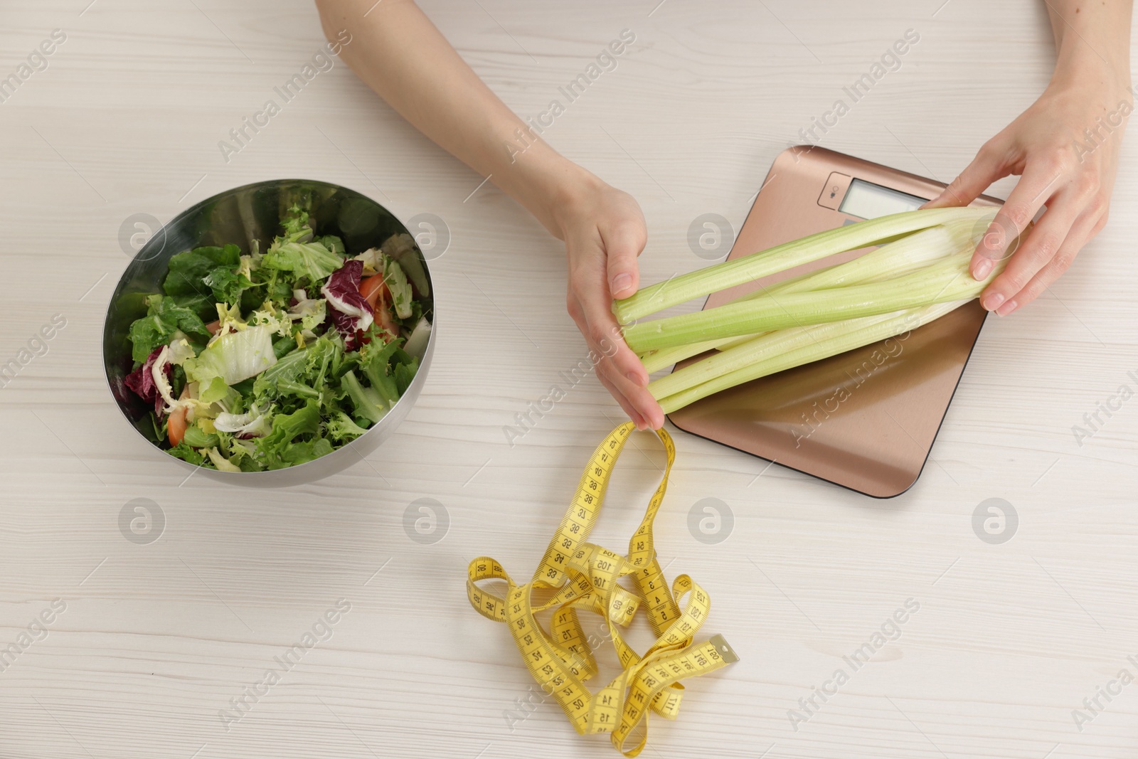 Photo of Woman weighting celery on kitchen scale at white wooden table, top view