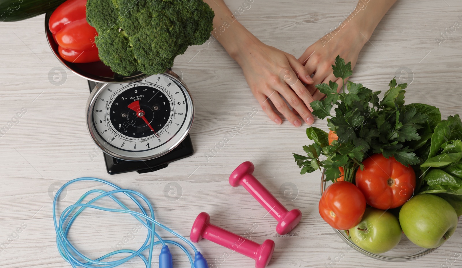 Photo of Woman at light wooden table with healthy food, kitchen scale, skipping rope and dumbbells, top view