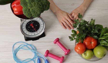 Photo of Woman at light wooden table with healthy food, kitchen scale, skipping rope and dumbbells, top view