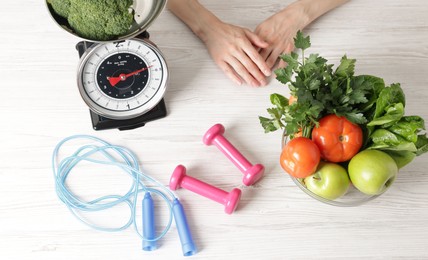 Photo of Woman at light wooden table with healthy food, kitchen scale, skipping rope and dumbbells, top view