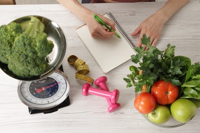 Photo of Woman developing diet plan at light wooden table with kitchen scale, measuring tape, dumbbells and healthy food, top view
