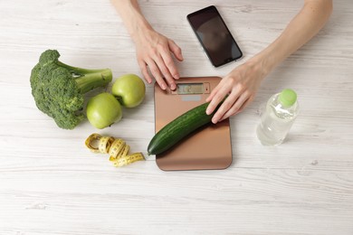 Photo of Woman weighting cucumber on kitchen scale at light wooden table, top view