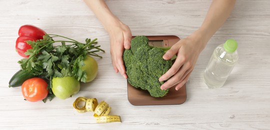 Photo of Woman weighting broccoli on kitchen scale at light wooden table, top view