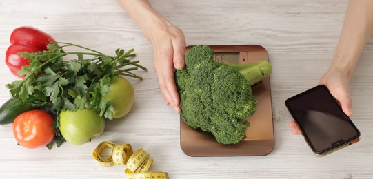 Photo of Woman with smartphone weighting broccoli on kitchen scale at light wooden table, top view
