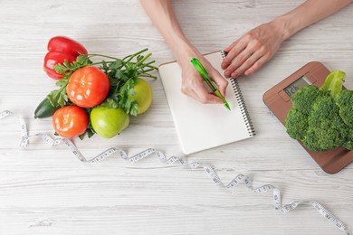 Photo of Woman developing diet plan at light wooden table with kitchen scale, measuring tape and healthy food, top view