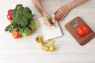 Photo of Woman developing diet plan at light wooden table with kitchen scale, measuring tape and healthy food, top view