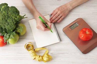 Photo of Woman developing diet plan at light wooden table with kitchen scale, measuring tape and healthy food, top view