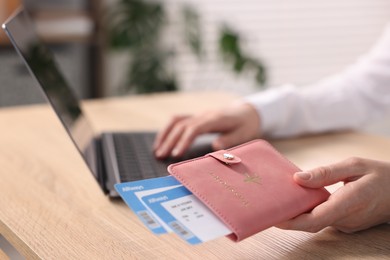 Photo of Woman with passport and flight tickets using laptop at wooden table indoors, closeup