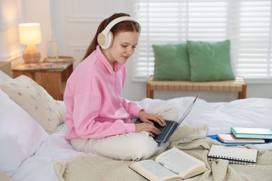 Photo of Teenage student in headphones using laptop while studying on bed at home