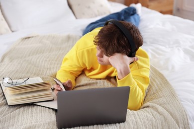Photo of Teenage student in headphones using smartphone while studying on bed at home