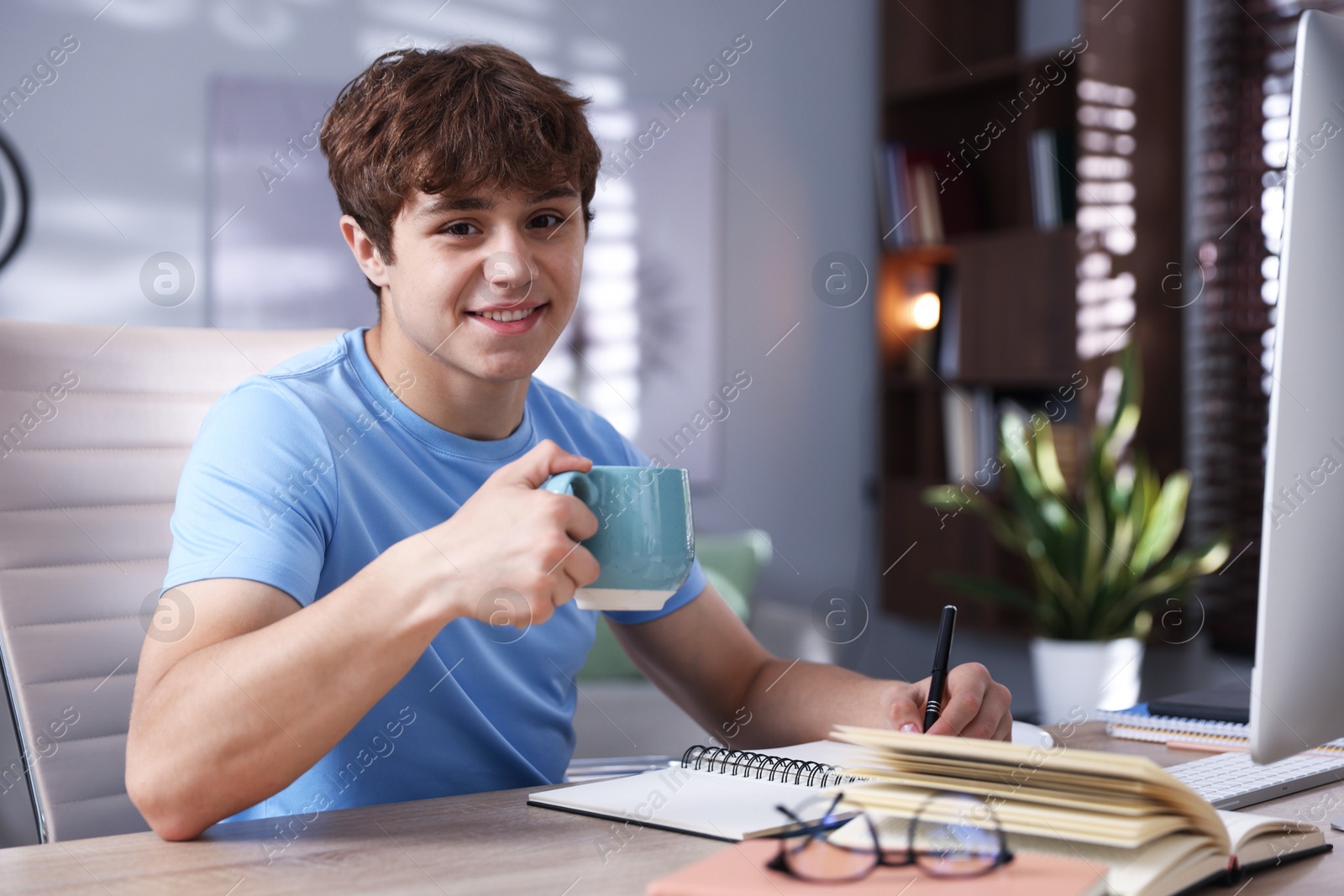 Teenage student with cup of drink taking notes while studying at table indoors Photo of Teenage student with cup of drink taking notes while studying at table indoors