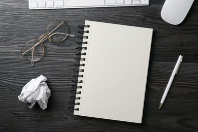 Photo of Notebook, pen, crumpled paper, glasses, computer keyboard and mouse on black wooden table, flat lay