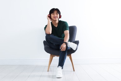 Photo of Handsome Asian man listening to music in headphones on armchair near white wall
