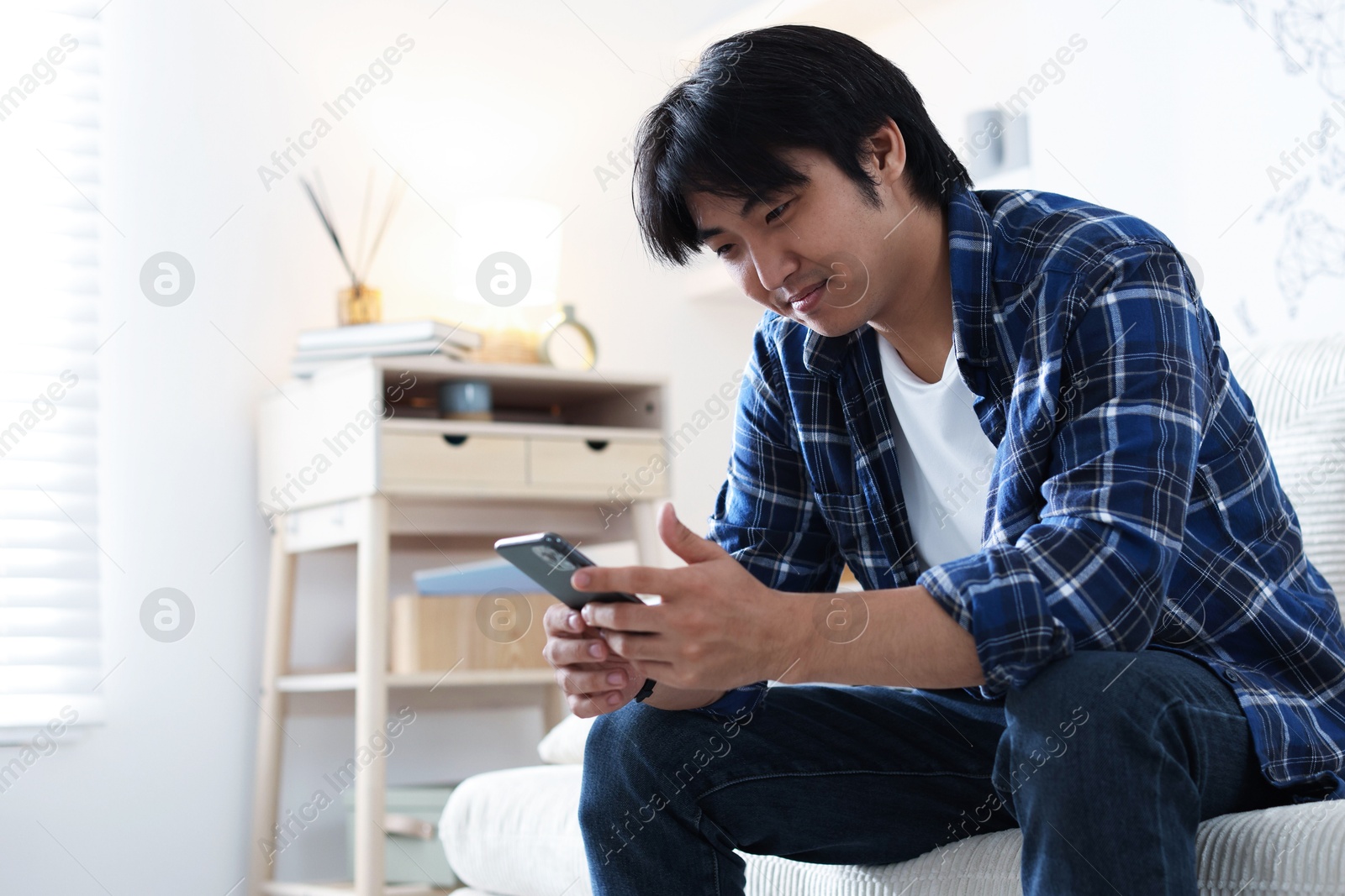 Handsome Asian man with smartphone on sofa at home. Space for text Photo of Handsome Asian man with smartphone on sofa at home. Space for text
