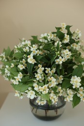 Photo of Beautiful jasmine flowers in vase on white table near beige wall indoors, closeup