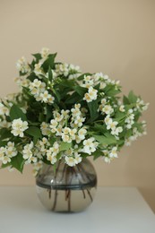 Photo of Beautiful jasmine flowers in vase on white table near beige wall indoors, closeup