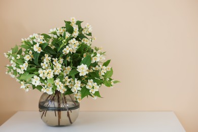 Photo of Beautiful jasmine flowers in vase on white table near beige wall indoors, space for text