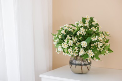 Photo of Beautiful jasmine flowers in vase on white table near beige wall indoors, space for text