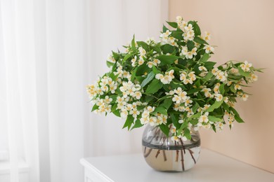 Photo of Beautiful jasmine flowers in vase on white table near beige wall indoors, space for text
