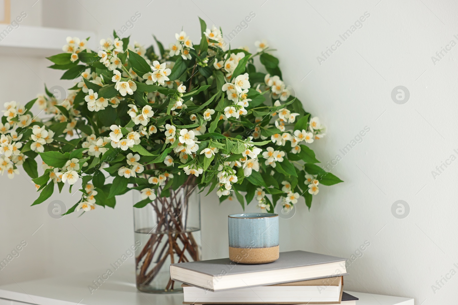 Beautiful jasmine flowers in vase, books and decor table near white wall indoors Photo of Beautiful jasmine flowers in vase, books and decor table near white wall indoors