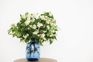 Photo of Beautiful jasmine flowers in vase on wooden table near white wall indoors. Space for text