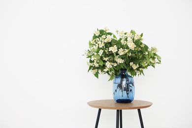 Photo of Beautiful jasmine flowers in vase on wooden table near white wall indoors, space for text