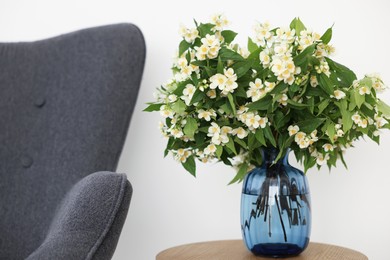 Photo of Beautiful jasmine flowers in vase on wooden table near armchair indoors