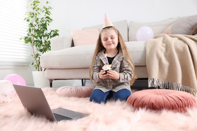 Photo of Birthday celebration. Little girl with cupcake having video call via laptop at home