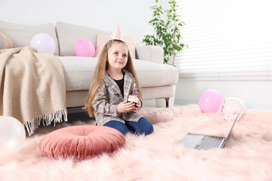 Photo of Birthday celebration. Little girl with cupcake having video call via laptop at home